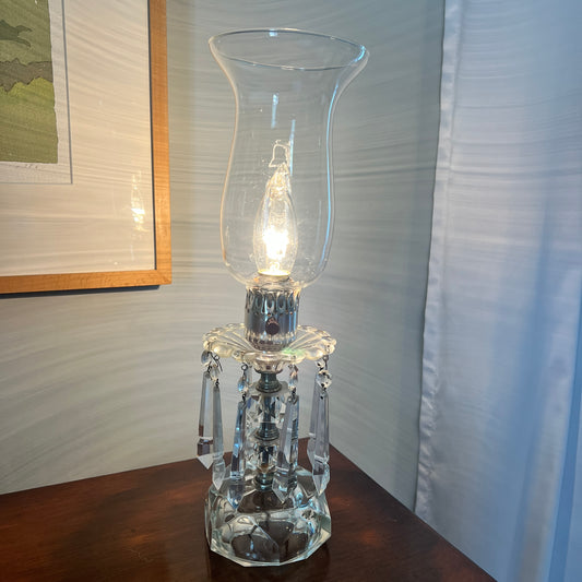 Clear glass hurricane lamp on a wooden surface with a light on inside, against a neutral wall.