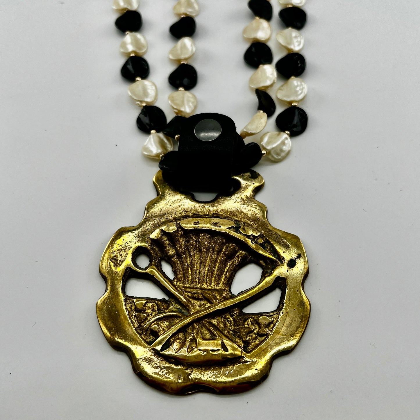 Vintage scythe and wheat horse brass medallion attached to triple-strand black and cream beaded necklace with leather connector.