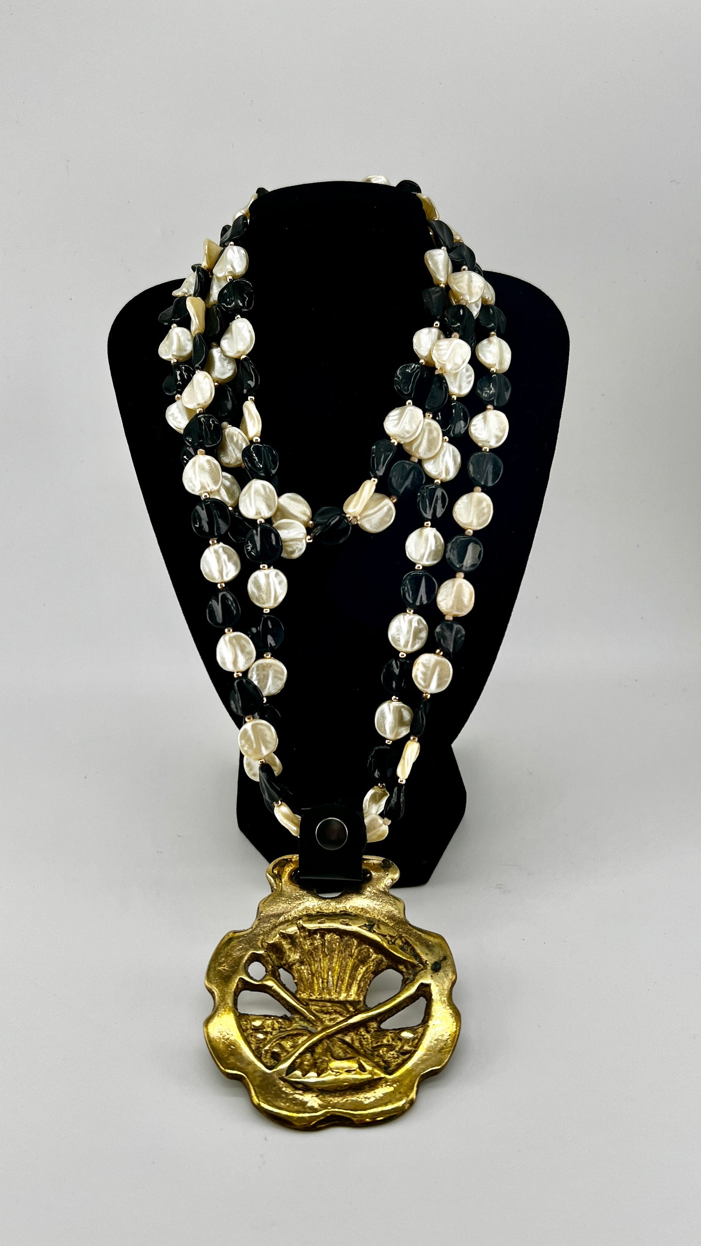 Vintage scythe and wheat horse brass medallion attached to triple-strand black and cream beaded necklace with leather connector.