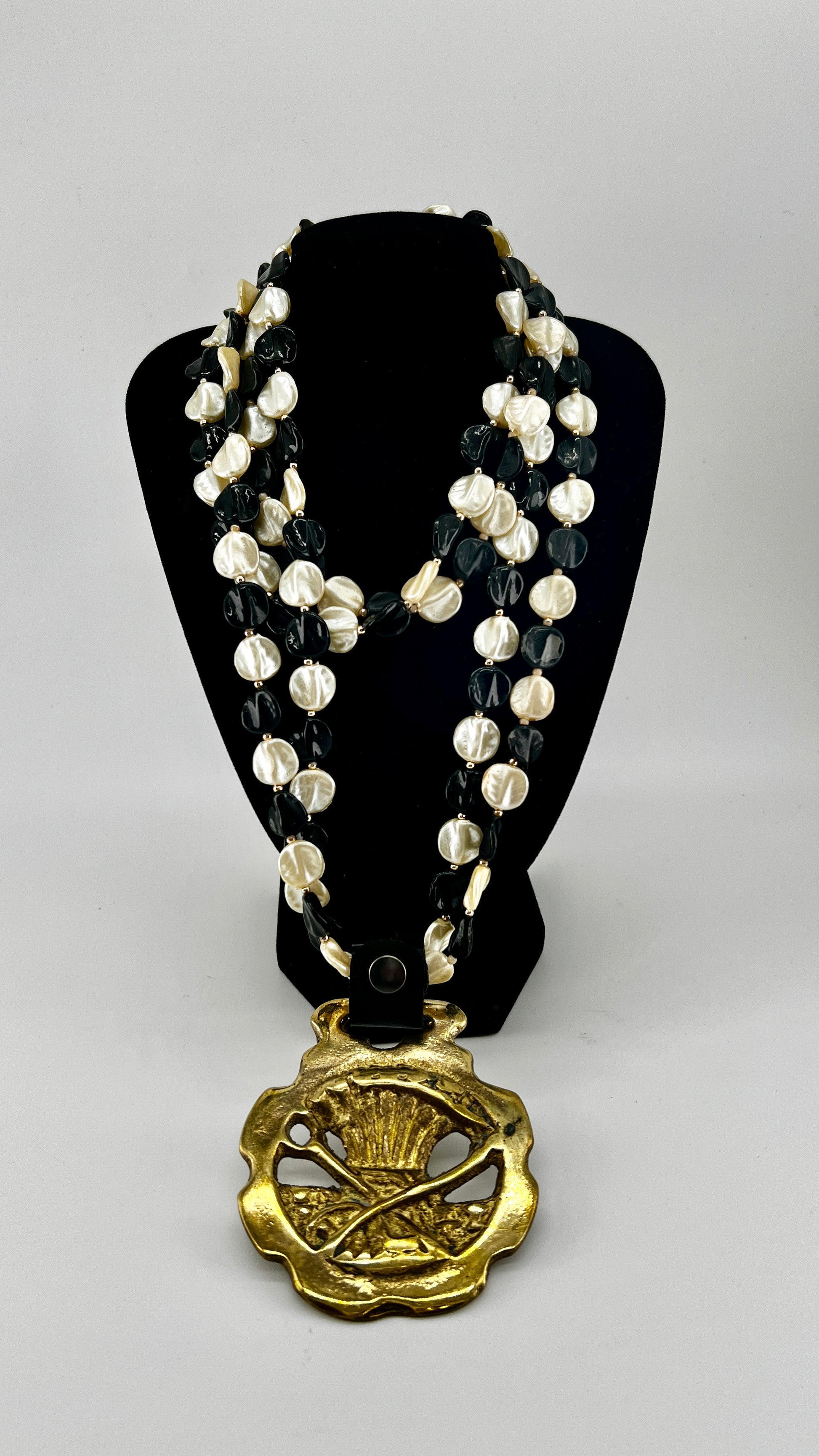 Vintage scythe and wheat horse brass medallion attached to triple-strand black and cream beaded necklace with leather connector.